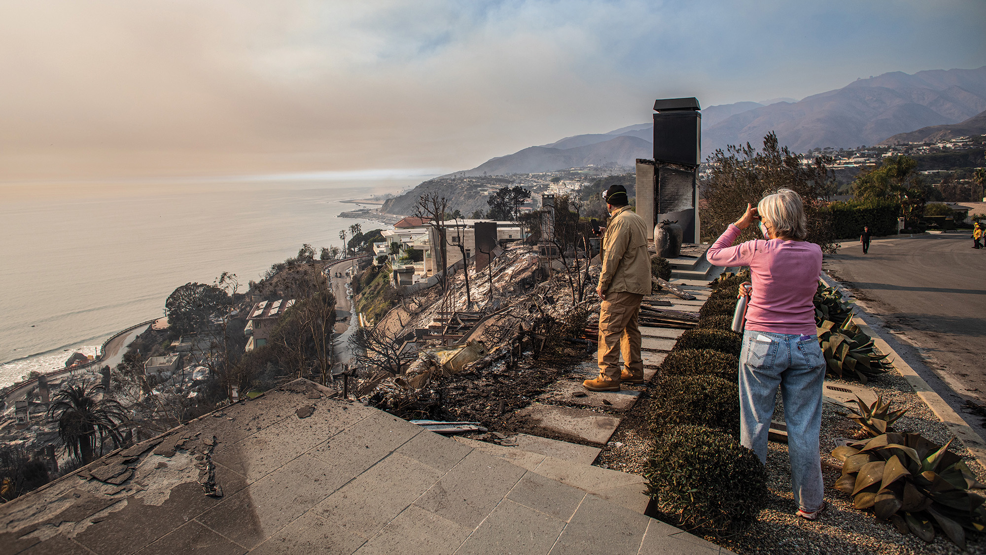 People surveying the damage of the Palisades Fire overlooking the Pacific Ocean with hills in the distance