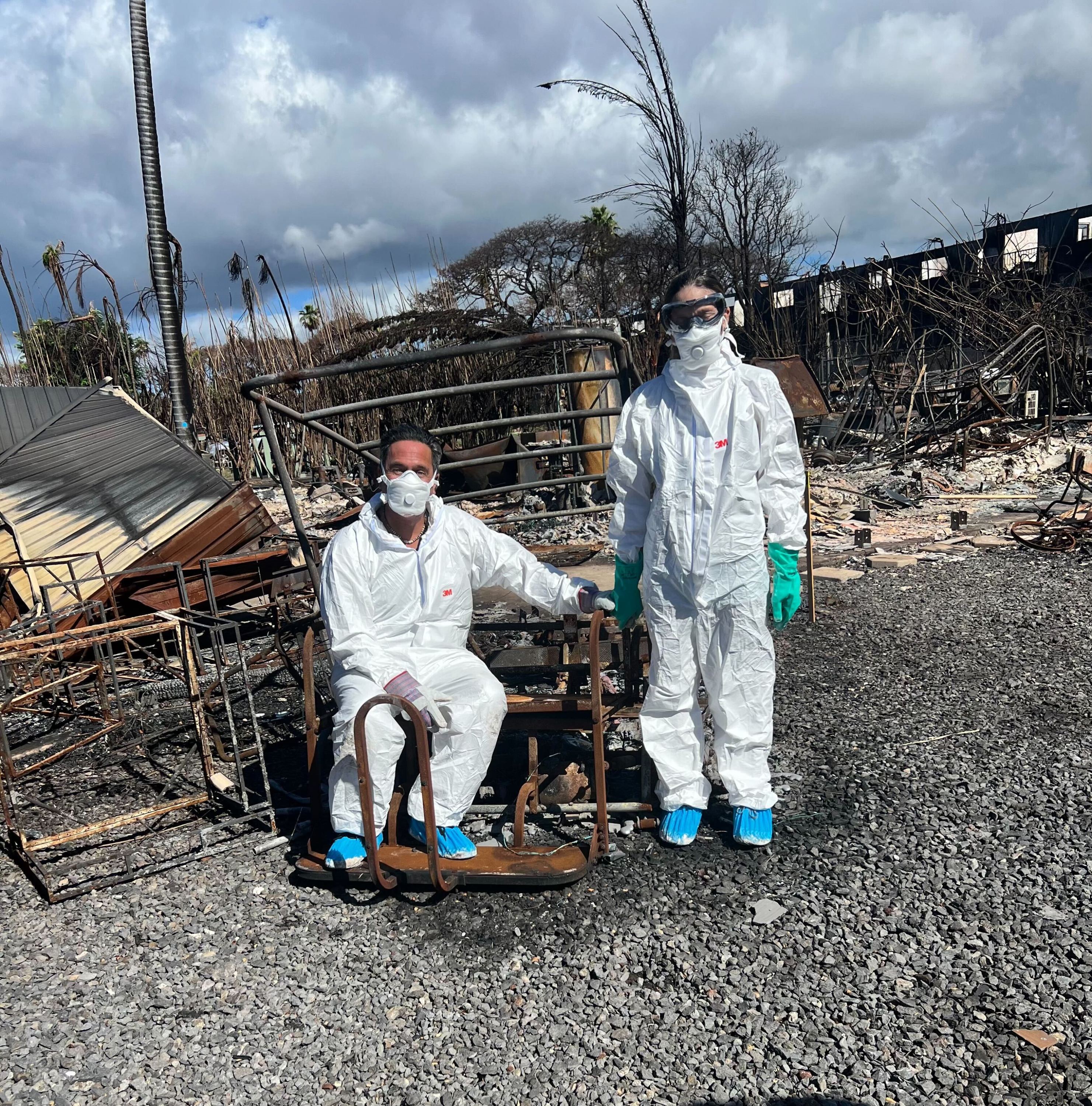 Two people stand in the ashes and rubble caused by the Lahaina, Hawaii fire while wearing full personal protective equipment suits.