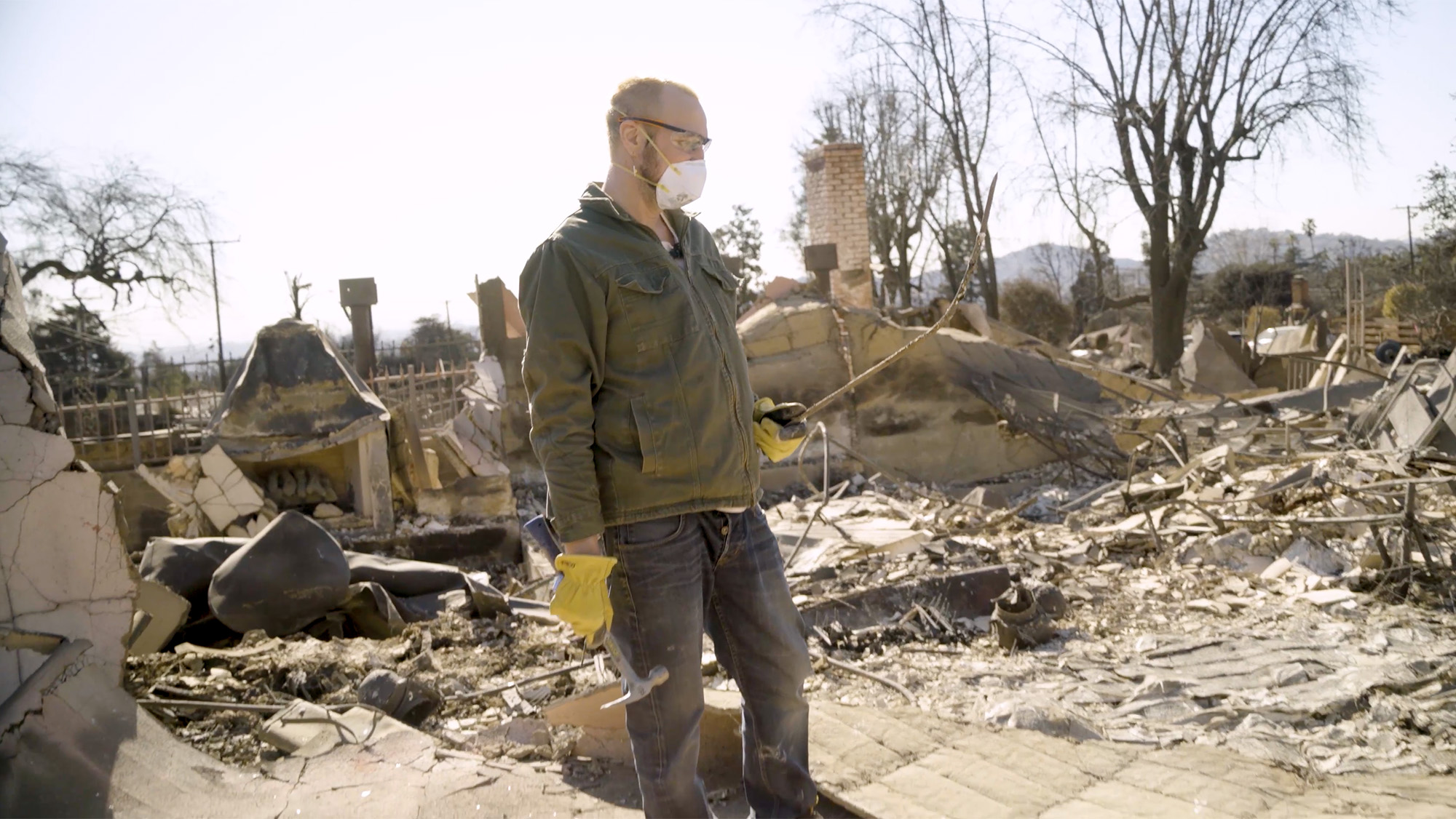 A man standing amid the ruins of his burned house, holding a long piece of metal